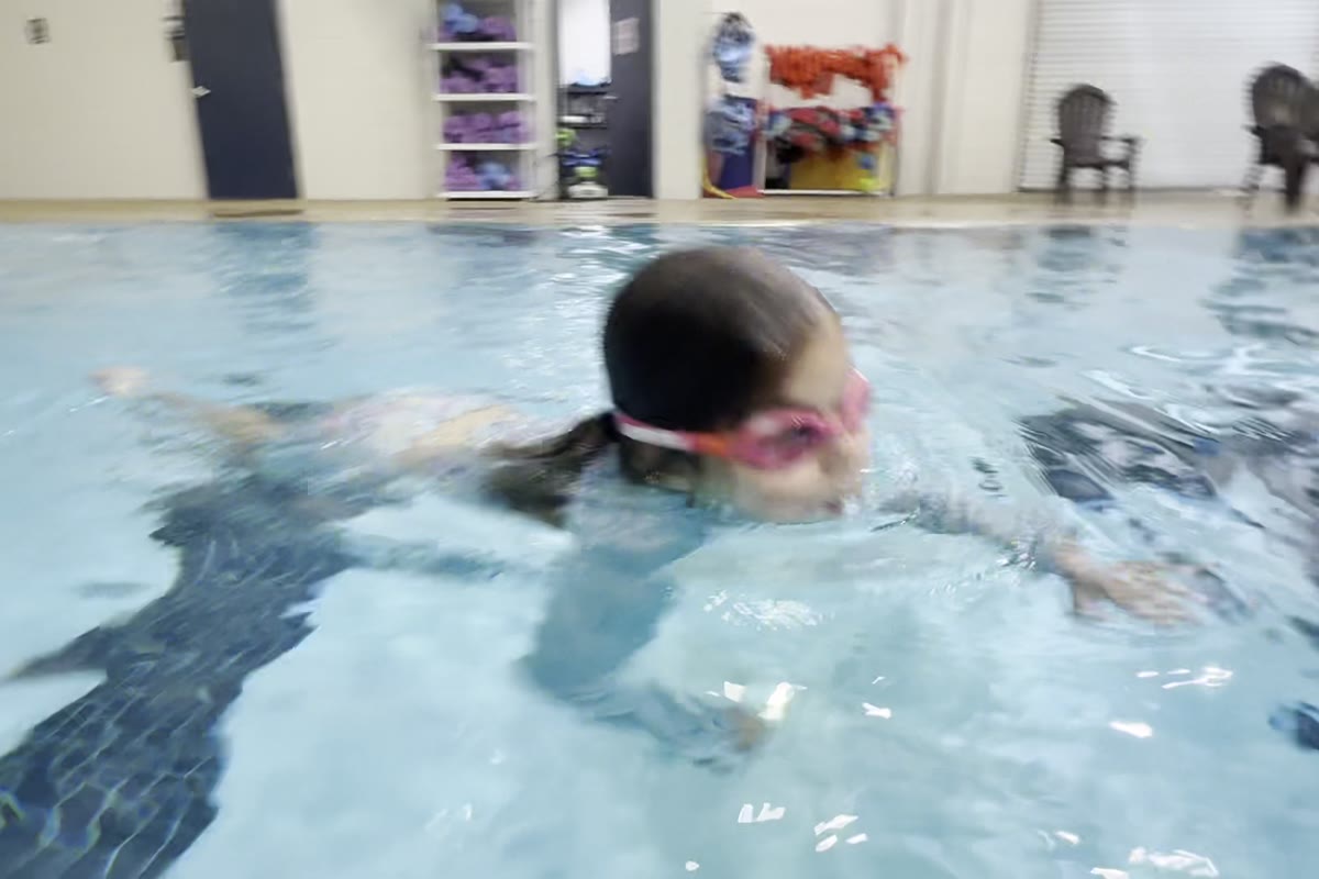 A young swimmer in pink goggles lifting her head to breathe as she swims toward the camera