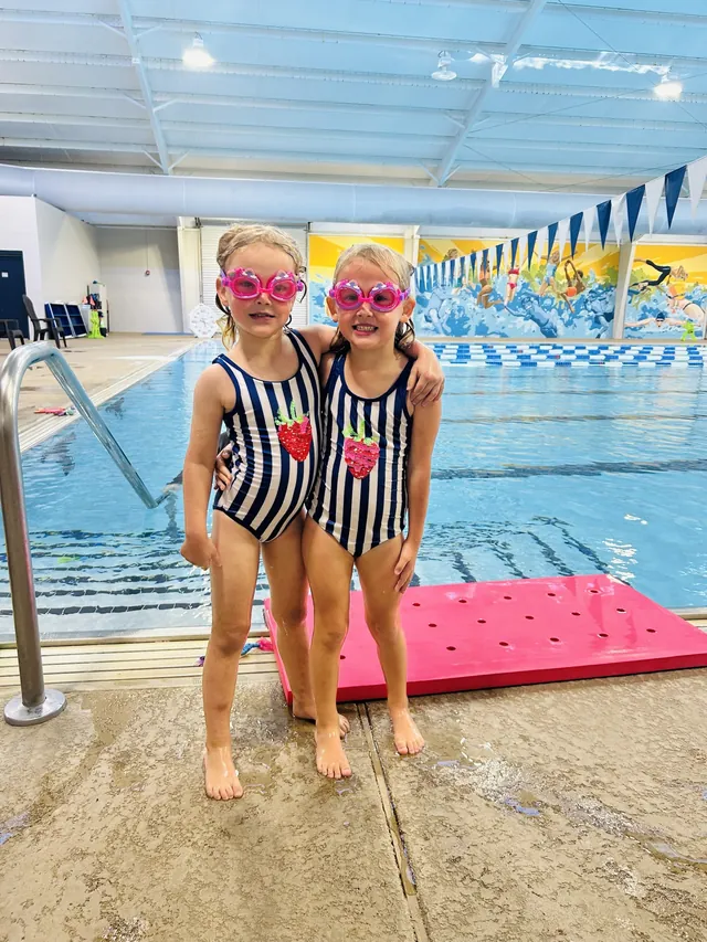 Two young swimmers in matching striped suits and pink goggles, arm in arm on the pool deck.